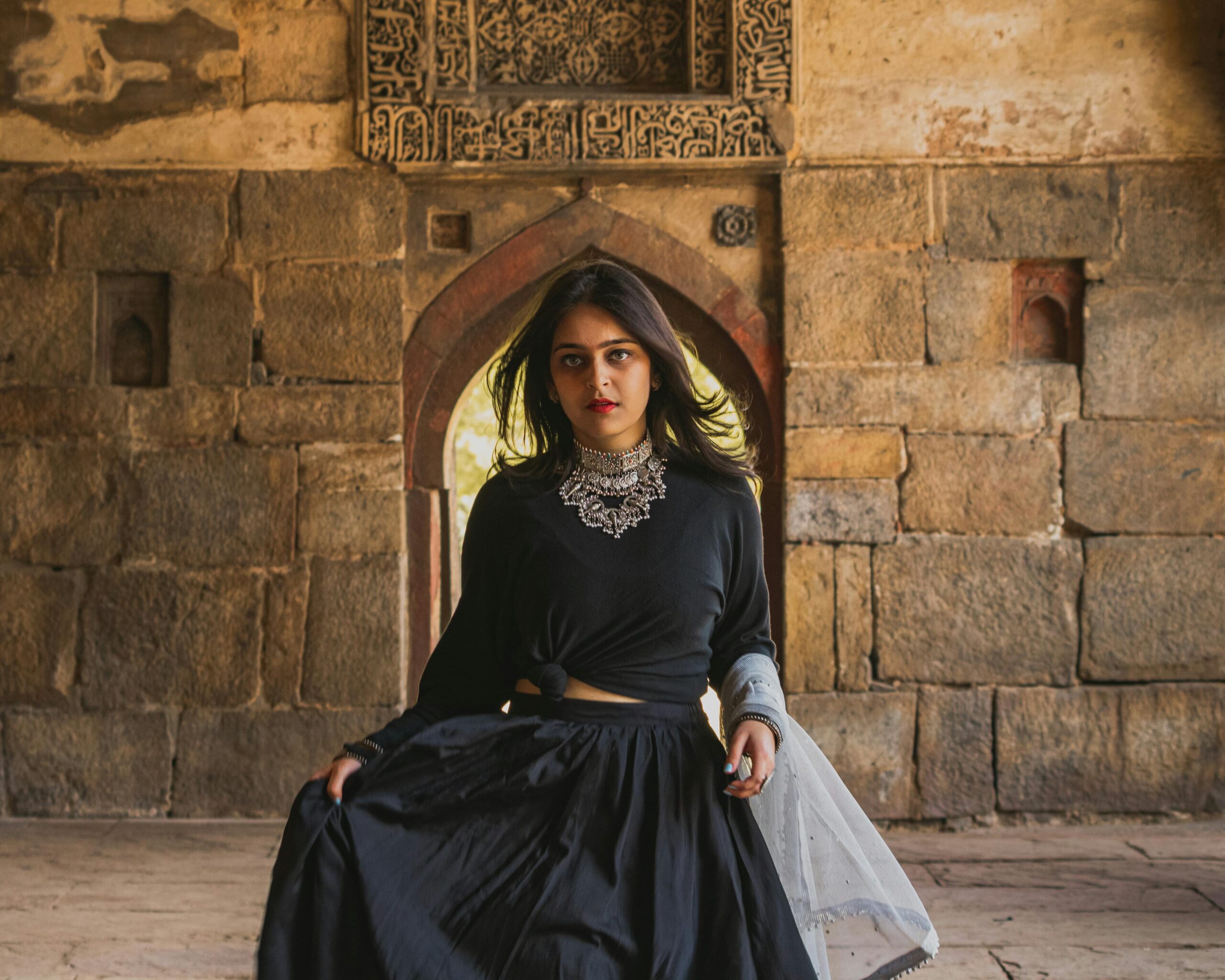 Woman in black outfit with jewelry walks through an ancient stone hallway.